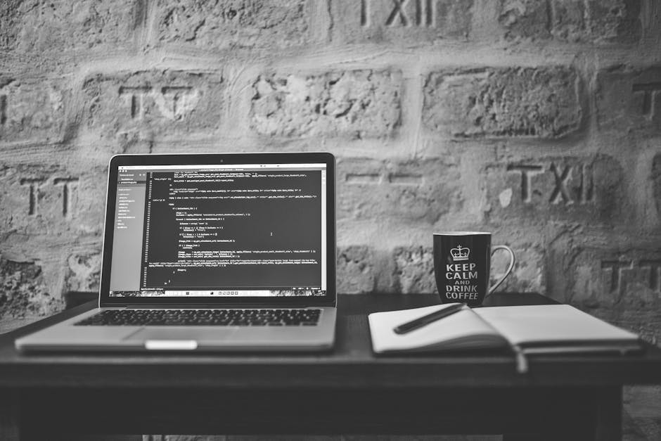 A black-and-white photograph of a workspace featuring an open laptop displaying lines of code on a wooden desk. To the right of the laptop, there is a white notebook with a black pen resting on top, a