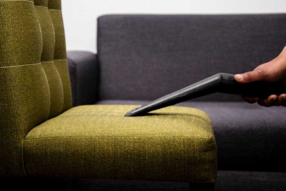 A person uses a handheld vacuum cleaner to vacuum the olive-green fabric upholstery of a modern armchair, positioned in a living room setting with a dark gray sofa in the background. The room is well-