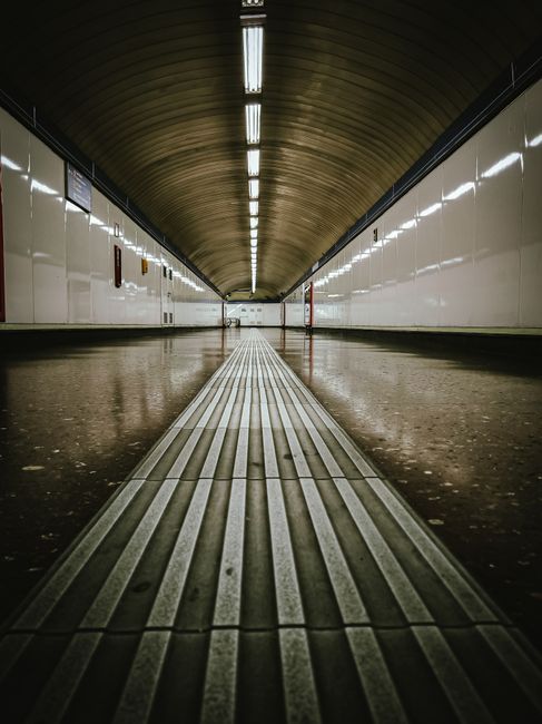 A person with a backpack is descending an escalator inside Streatham station. The station features modern architectural elements, including metal support beams, glass panels, and concrete walls. The e